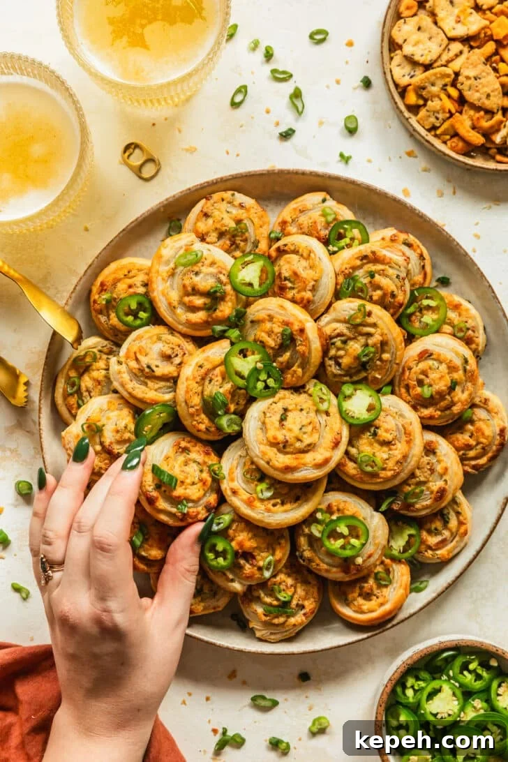 A hand reaching for a pinwheel on a beige plate of baked jalapeño popper pinwheel next to glasses of beer, an orange linen, and brown bowls of peppers and snack mix with a tan background.