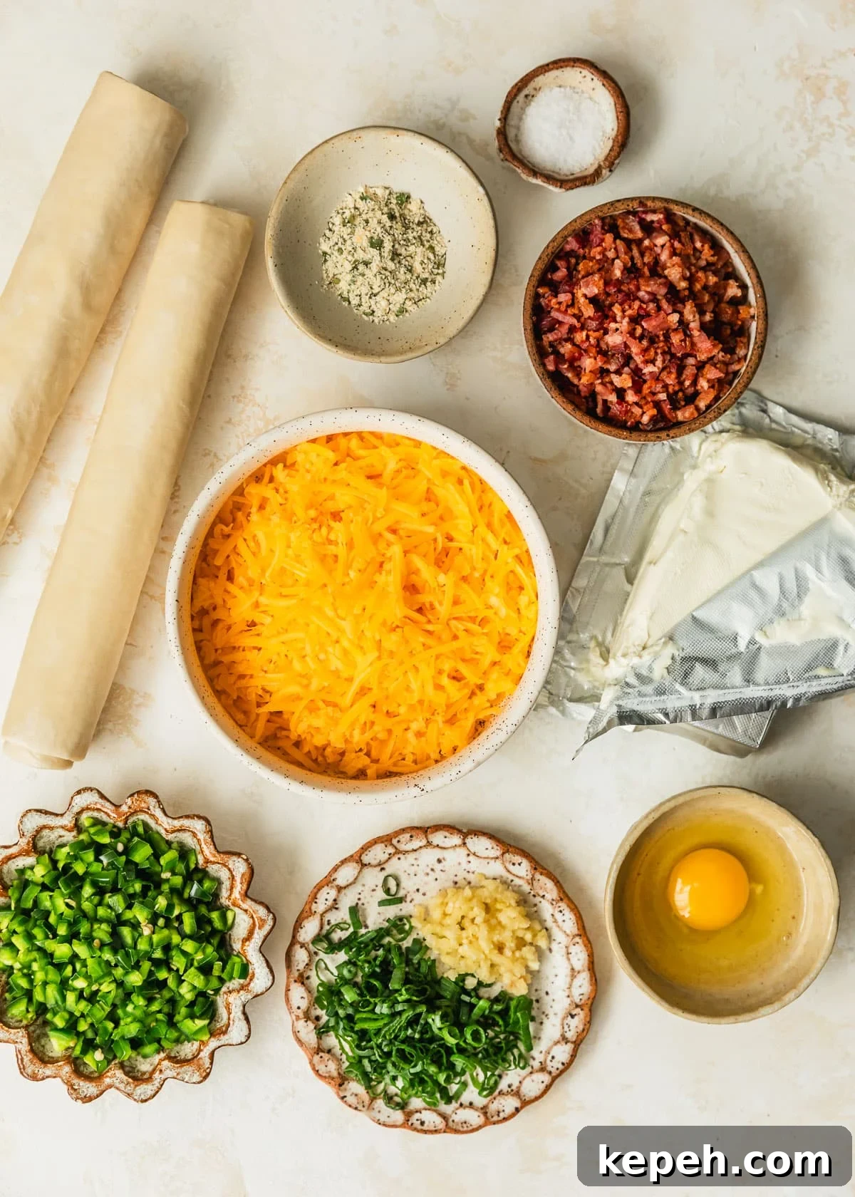 White bowls of cheddar cheese, bacon, ranch seasoning, salt, jalapeños, scallions, garlic, and an egg on a tan counter next to a block of cream cheese and puff pastry.