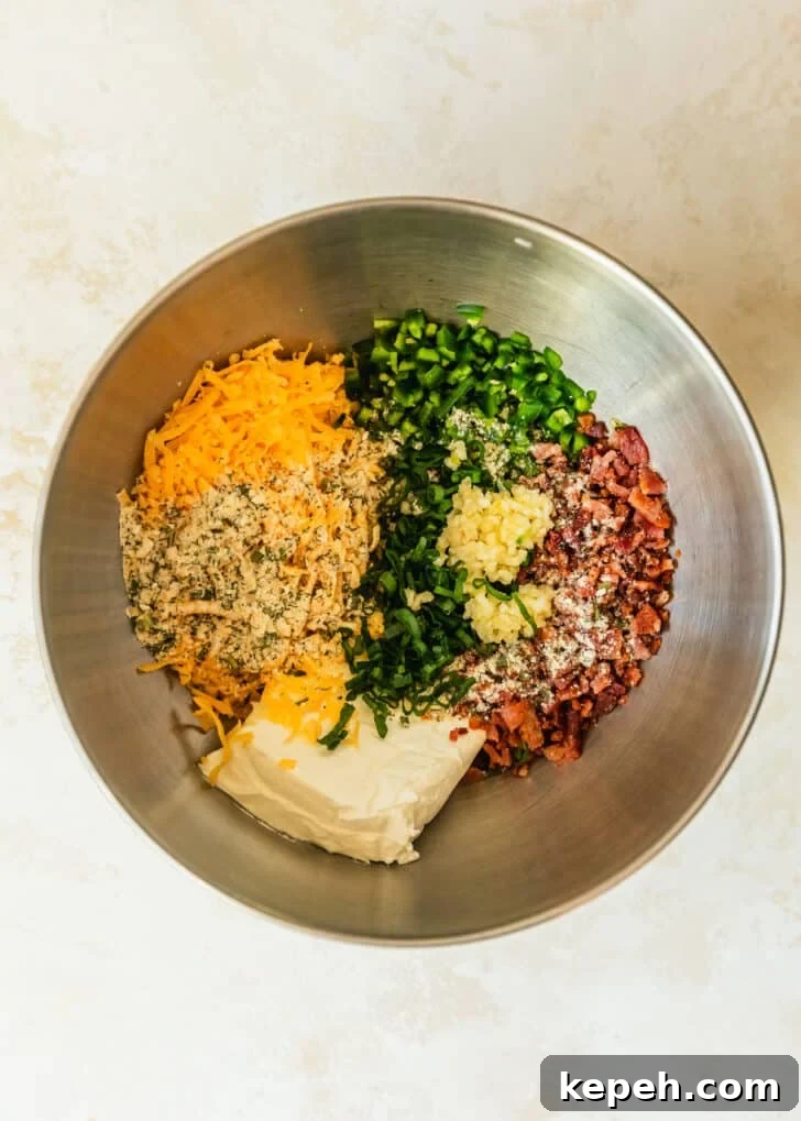 A silver bowl of cream cheese, cheddar, peppers, bacon, garlic, green onions, and ranch seasoning on a tan counter.