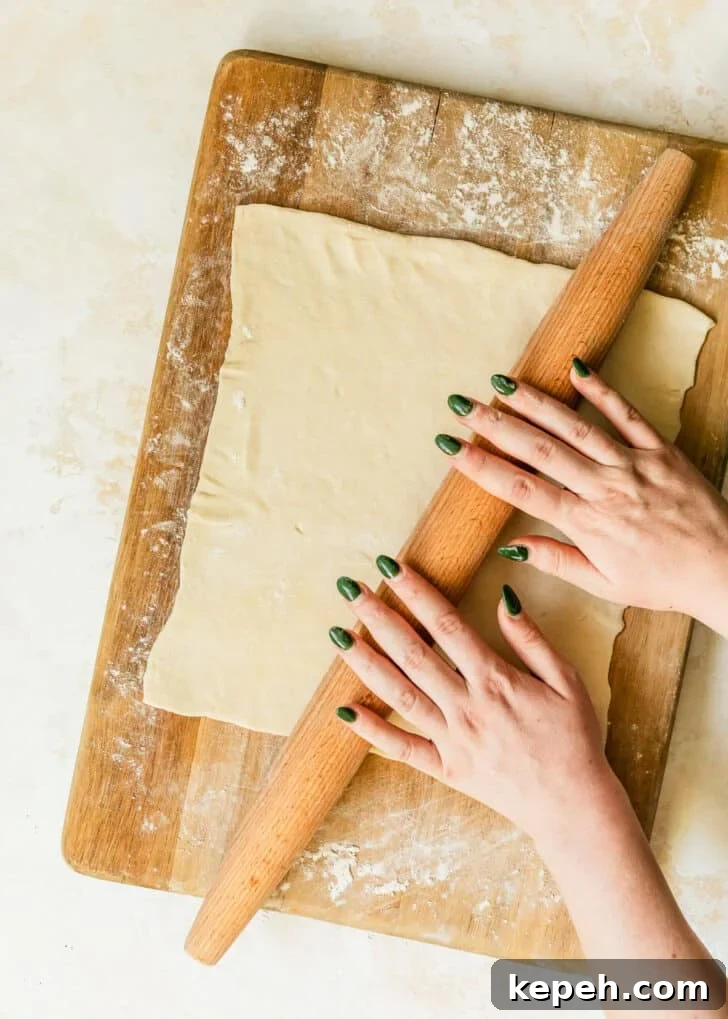 Hands using a rolling pin to roll puff pastry on a wood board.