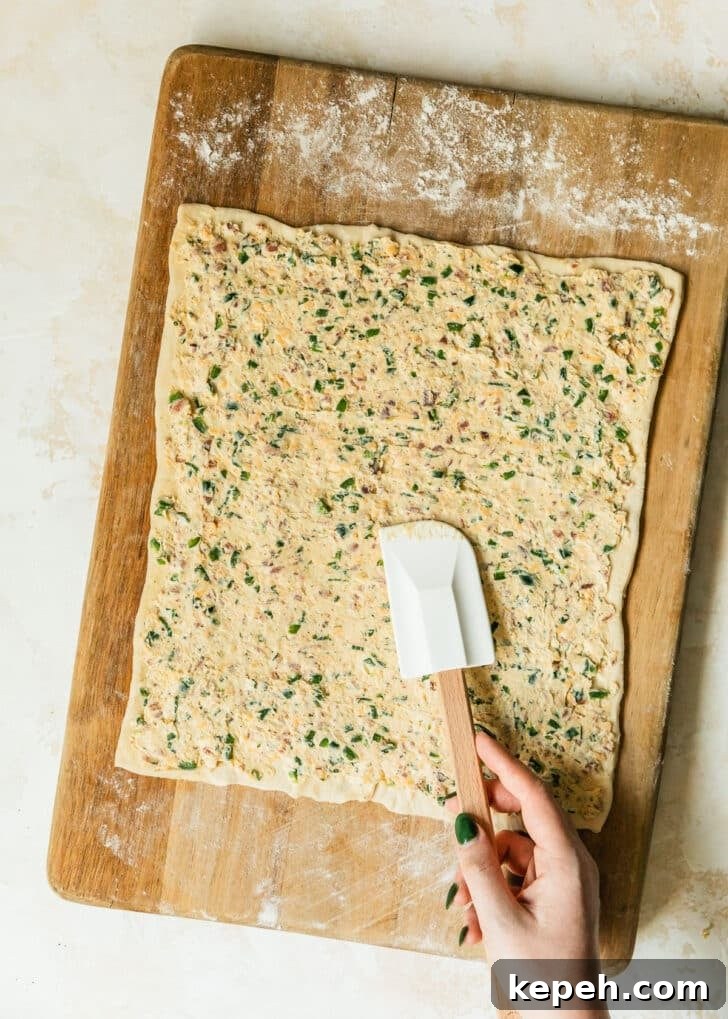 Hands using a rubber spatula to spread jalapeño cream cheese filling on puff pastry.