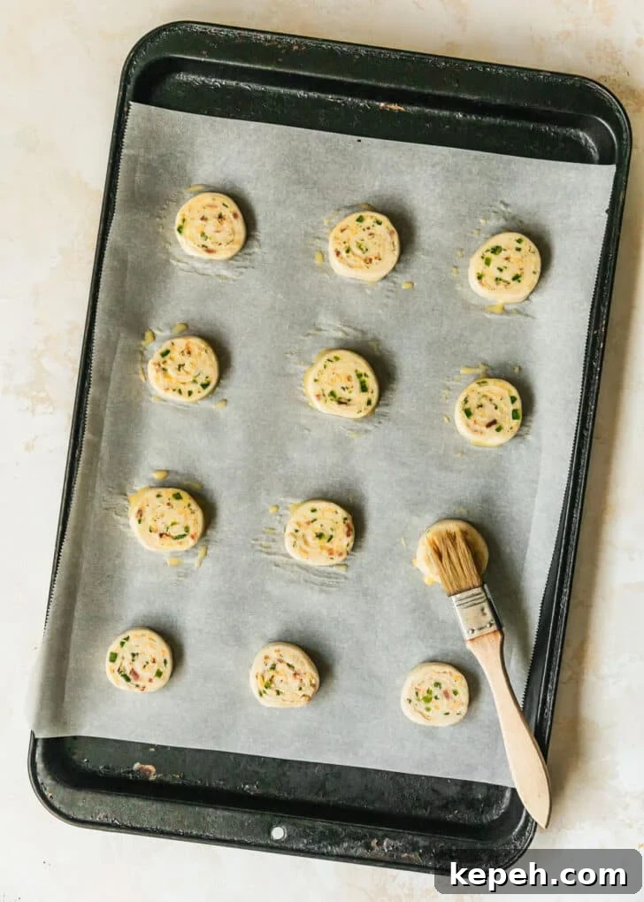Rows of jalapeño popper pinwheels with egg wash on a black sheet pan.