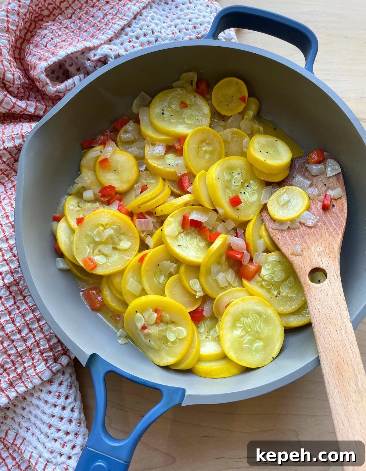 A blue and gray frying pan with buttery yellow summer squash and onions in it with a wooden spoon.