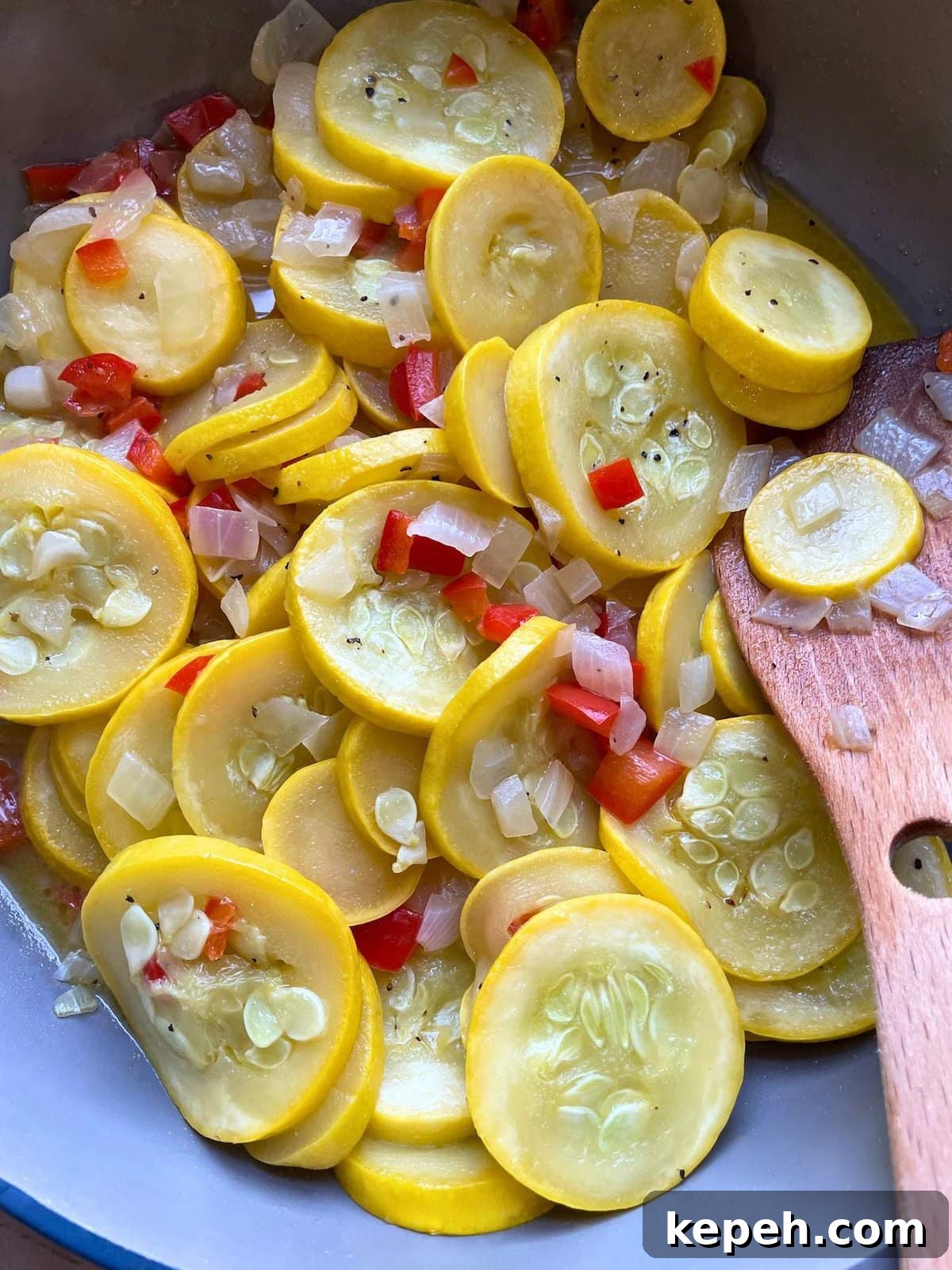 A blue and gray frying pan with sautéed squash, red bell peppers, and onions in it with a wooden spoon.