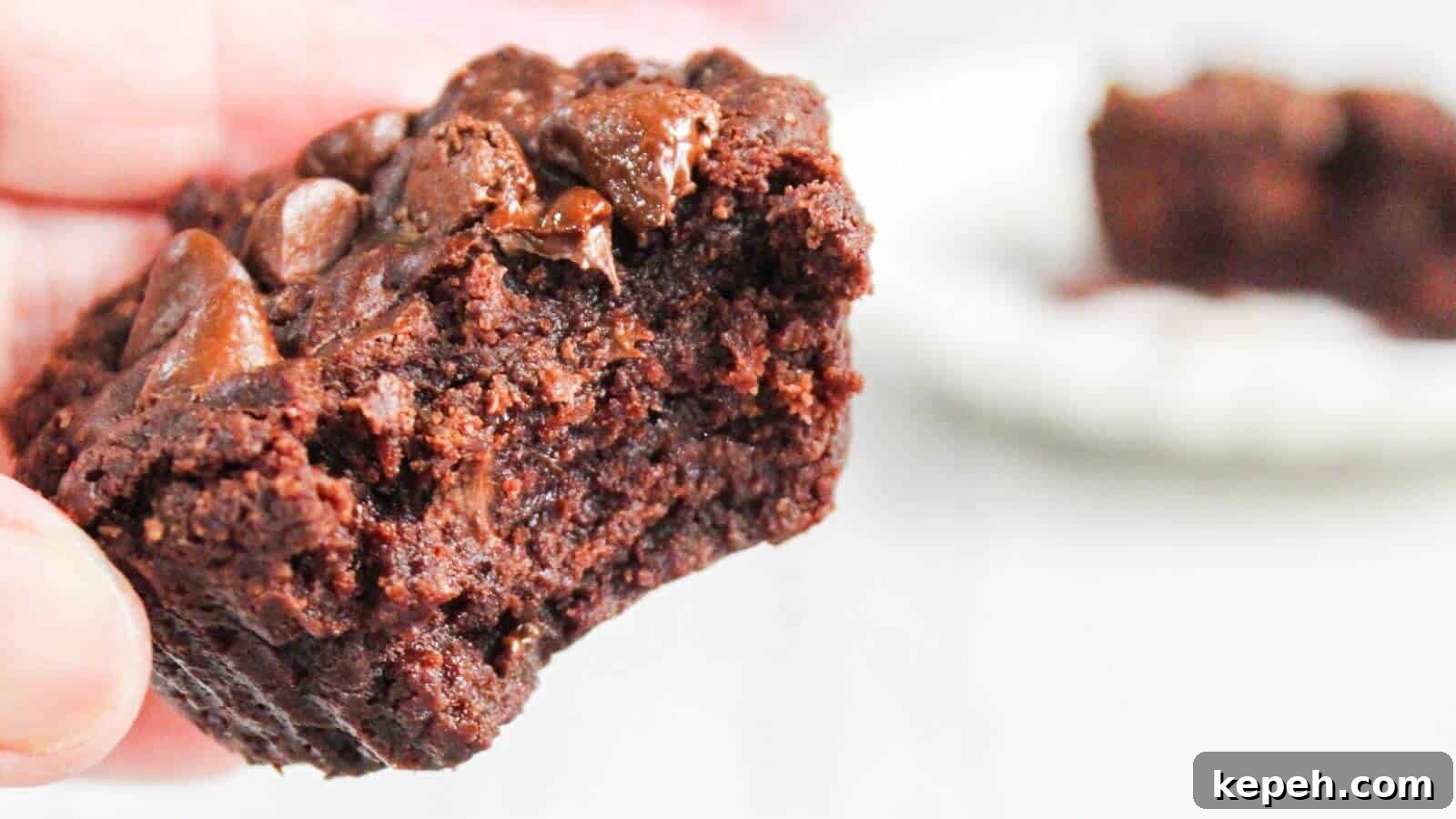 Close-up of a hand holding a half-eaten chocolate brownie with gooey chocolate chunks, with another partial brownie blurred in the background.