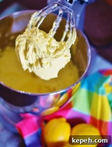 Lemon Buttercream Icing on a whisk attachment for a stand mixer. In the foreground are 3 lemons and a brightly colored pastel checked napkin.