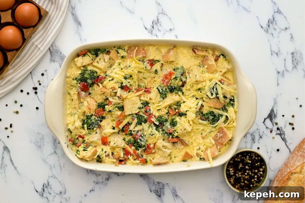 Bread, eggs, spinach, and roasted red peppers in baking dish before baking breakfast strata.