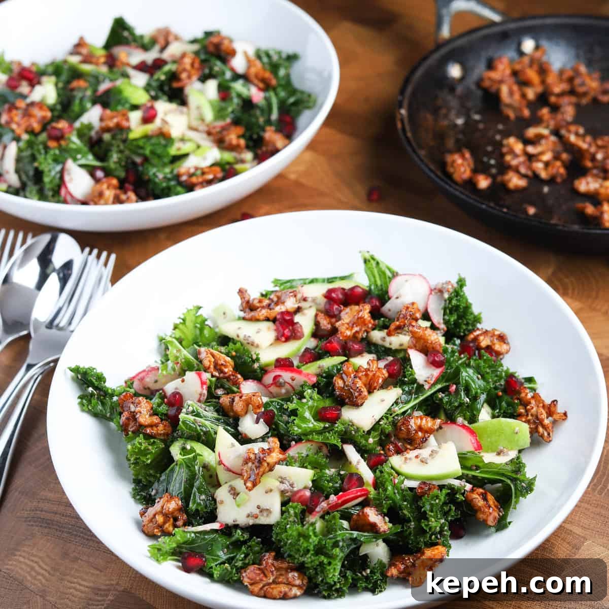 Square photo of two bowls of kale and apple salad with a small pan of caramelised walnuts in the background.