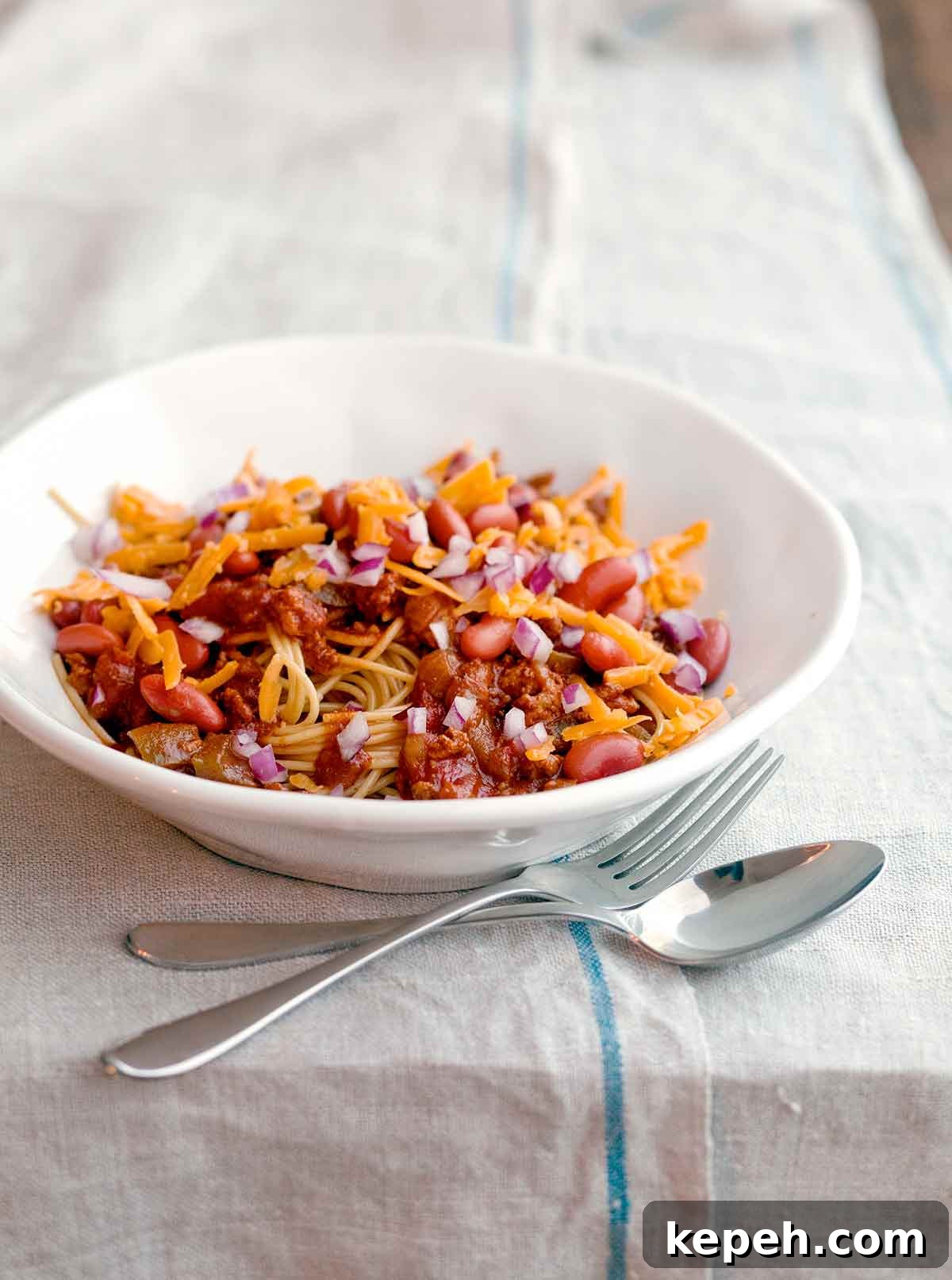 A white bowl of turkey Cincinnati chili with a fork and spoon resting beside it.