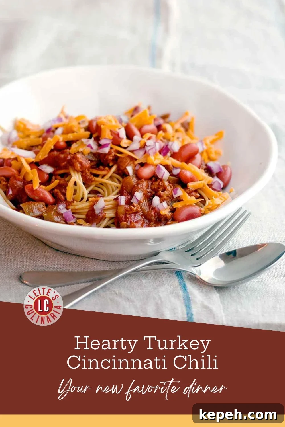 A white bowl of turkey Cincinnati chili with shredded cheese and diced red onion over spaghetti, with a fork and spoon on the table.