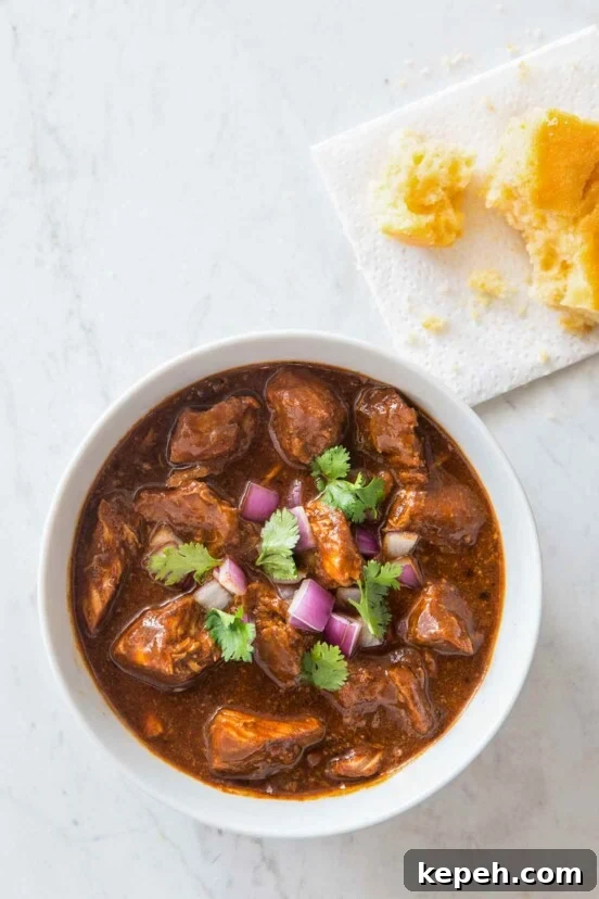 A white bowl filled with New Mexican red pork chili, garnished with red onion and cilantro.