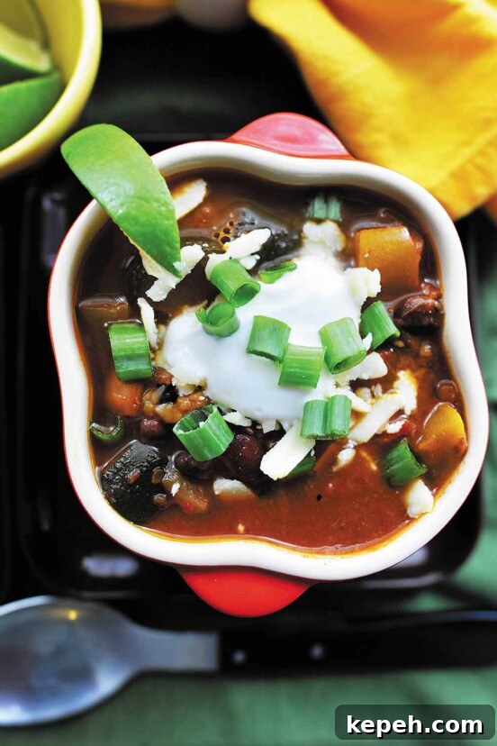 A white bowl with a slice of lime on the rim, filled with smoky chipotle vegetarian chili, sour cream, and topped with green onions.