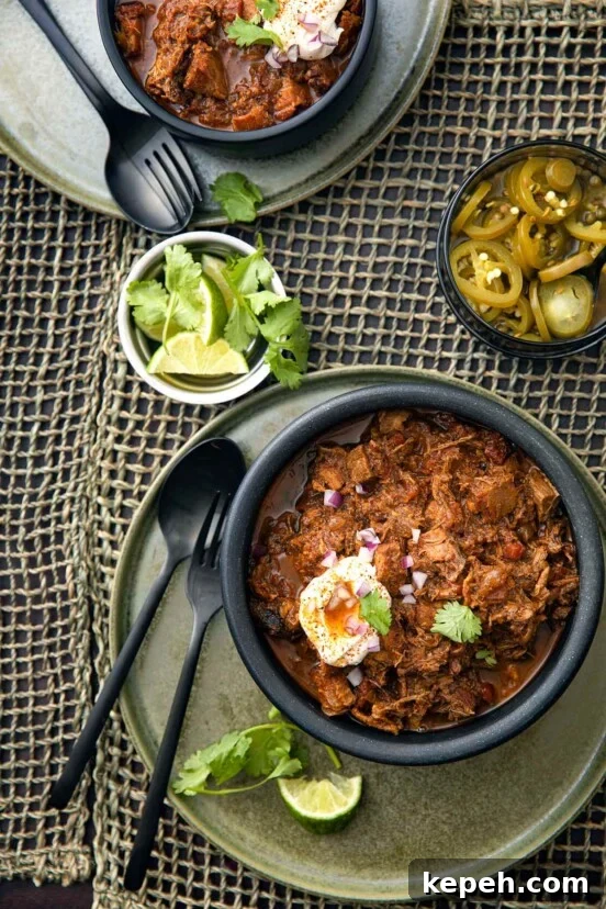 A bowl of Texas-style chili with pork and brisket with a fork, spoon, and bowls of lime, cilantro, and pickled jalapenos on the side.