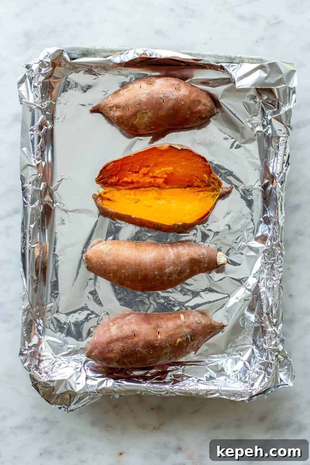 Baked sweet potatoes on a foil-lined sheet tray, with one of the sweet potatoes split open