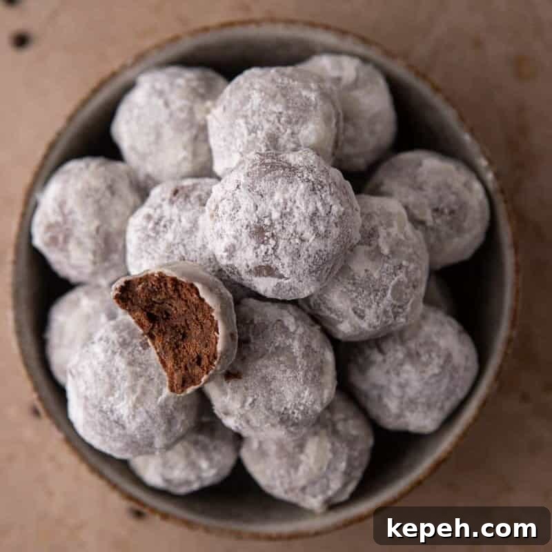 Rich Chocolate Snowball Cookies — Soft, Decadent Cocoa Bites 2 Bowl of powdered sugar–coated chocolate cookies with one cookie broken open.