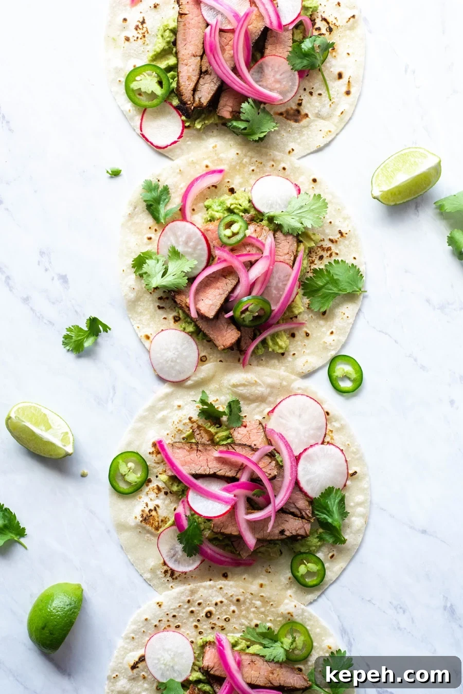 Marinated Steak & Avocado Tacos (Gluten-Free) 3 Multiple steak tacos lined up from the top to the bottom of the image on a white background with cilantro, thinly sliced jalapeno, pickled red onions
