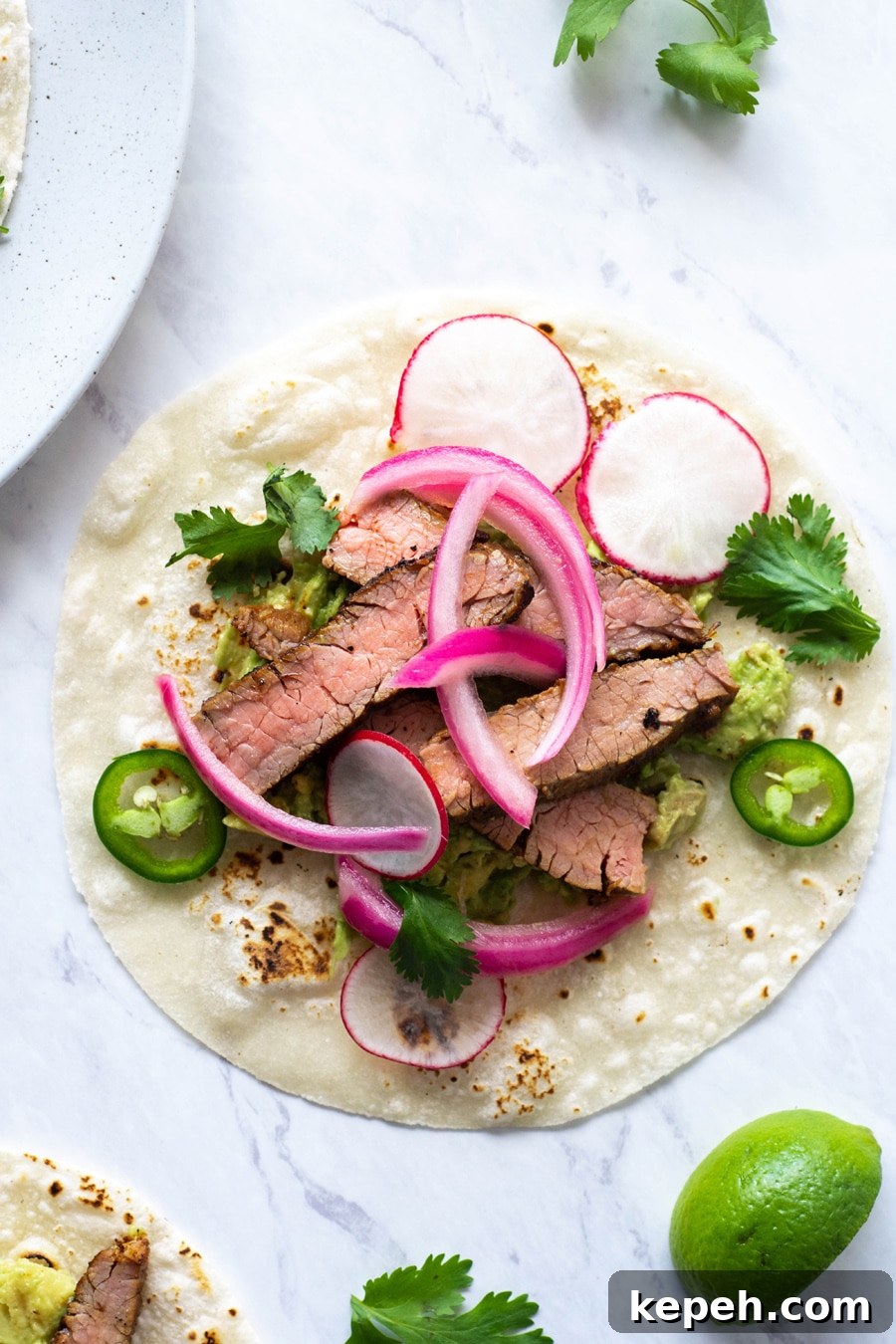 Marinated Steak & Avocado Tacos (Gluten-Free) 4 A close up shot of a steak taco on a white background with cilantro, thinly sliced jalapeno, pickled red onions
