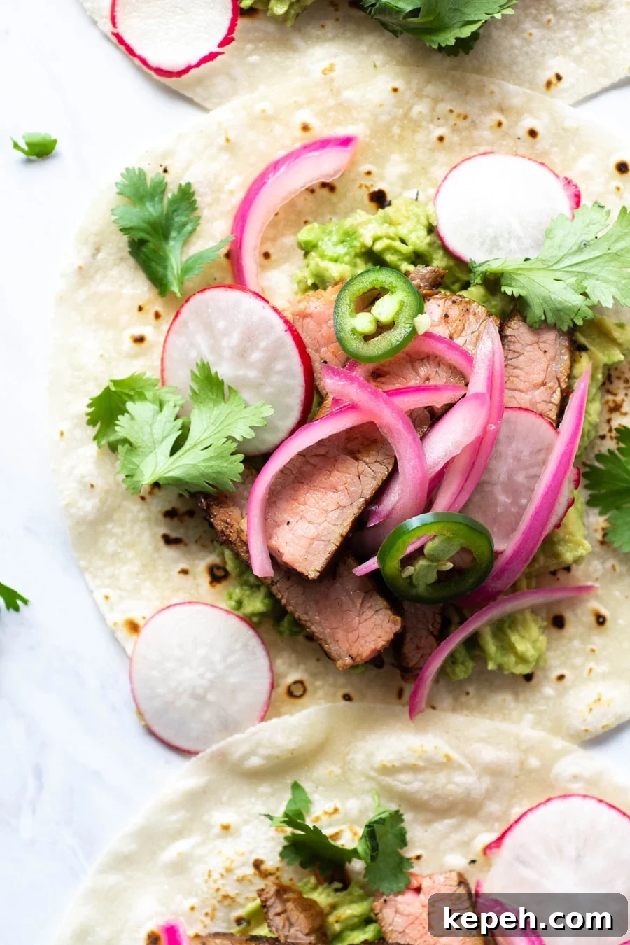 Marinated Steak & Avocado Tacos (Gluten-Free) 5 Close up shot of a steak taco on a white background with cilantro, thinly sliced jalapeno, pickled red onions