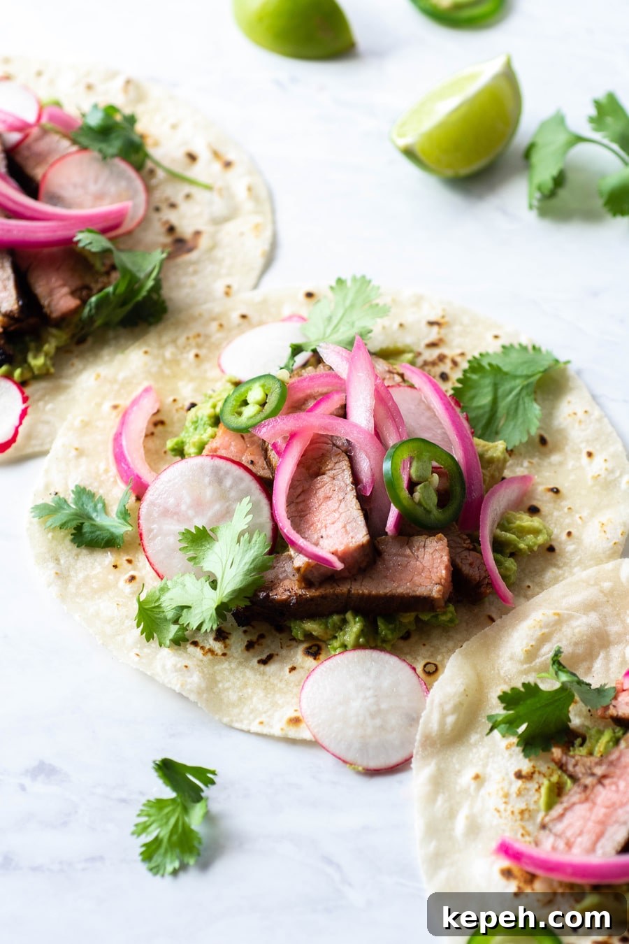 Marinated Steak & Avocado Tacos (Gluten-Free) 7 Side angle of multiple steak tacos on a white background with cilantro, thinly sliced jalapeno, pickled red onions