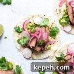 Multiple steak tacos on a white background with cilantro, thinly sliced jalapeno, pickled red onions