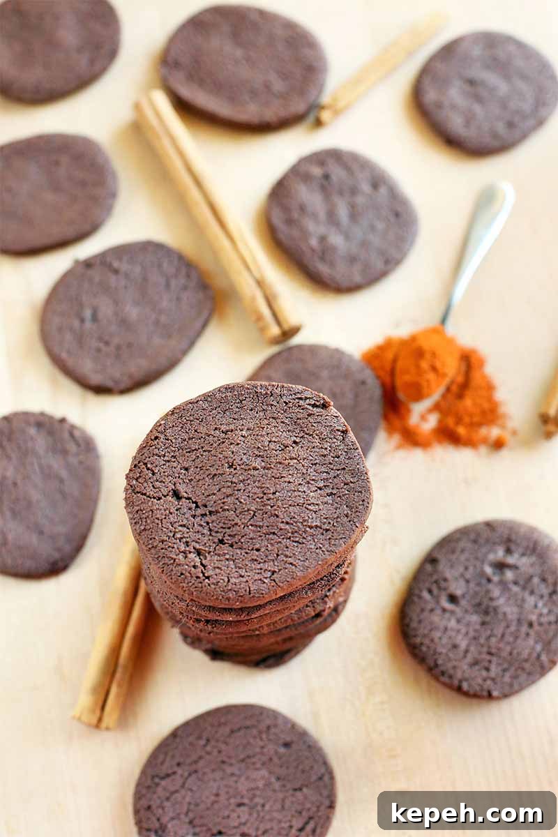 Spicy Mexican Hot Chocolate Cookies to Kick Off Baking Season 3 Top-down vertical shot of a tall stack and scattered chocolate cookies, with whole brown cinnamon sticks and an overfilled spoonful of orange cayenne pepper powder, on a beige surface.
