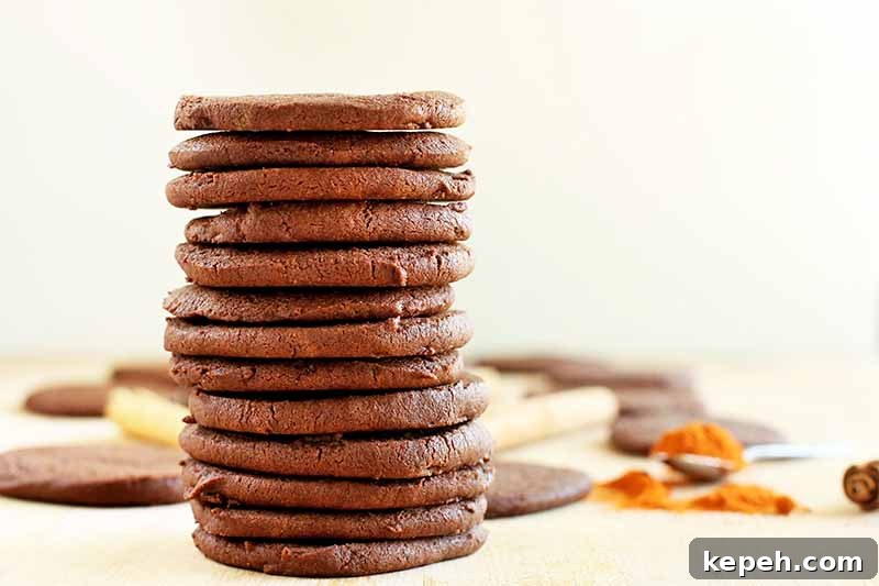 Spicy Mexican Hot Chocolate Cookies to Kick Off Baking Season 9 A stack of 13 chocolate cookies in the foreground, with more of the baked treats, cinnamon sticks, and a spilled spoonful of cayenne pepper in soft focus in the background, on a beige surface against an off-white wall.