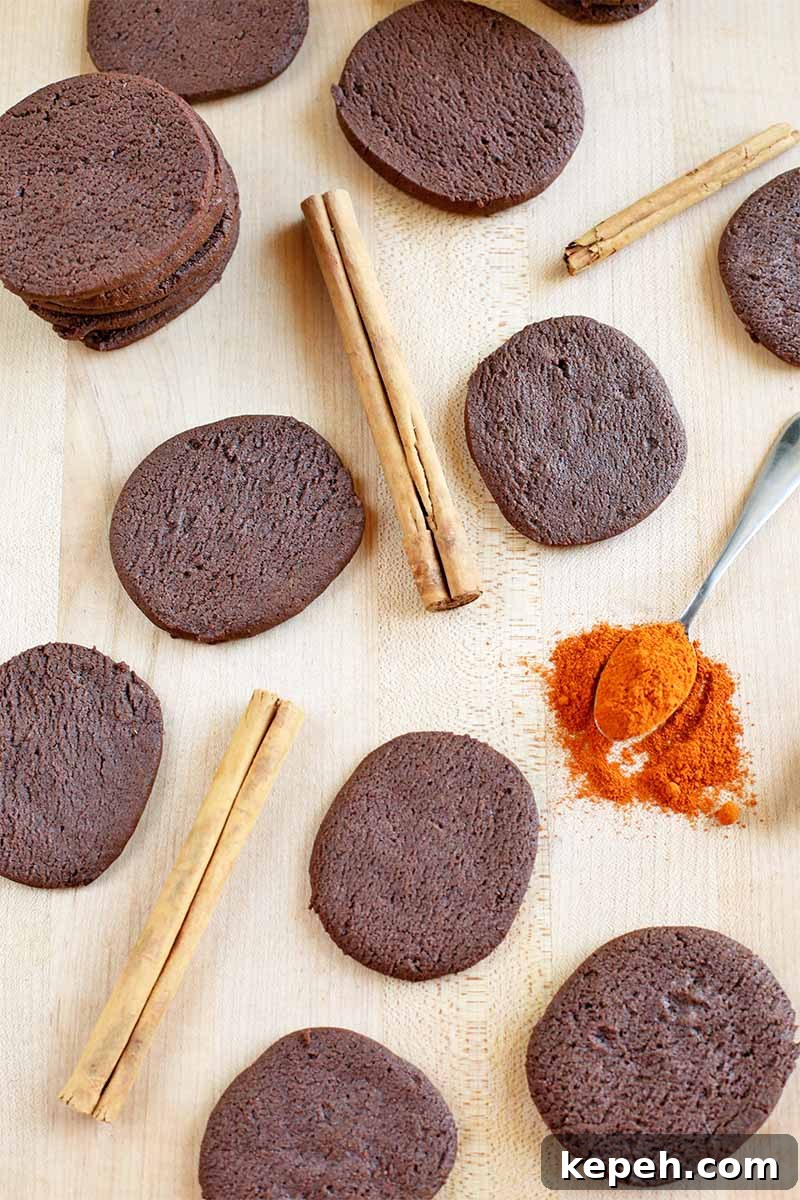 Spicy Mexican Hot Chocolate Cookies to Kick Off Baking Season 4 Top-down shot of chocolate cookies, cinnamon sticks, and a spoonful of orange cayenne pepper that has spilled onto the beige countertop.