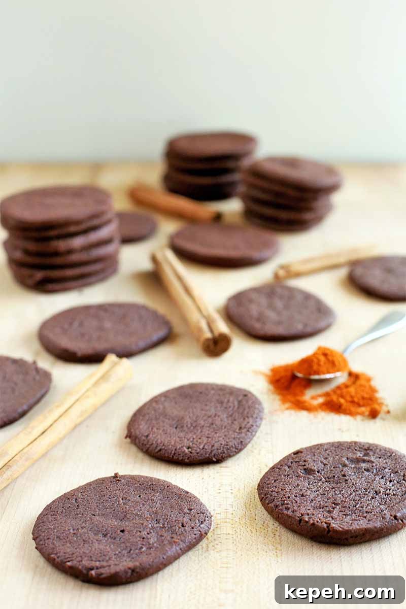 Spicy Mexican Hot Chocolate Cookies to Kick Off Baking Season 5 Vertical image of chocolate cookies, cinnamon sticks, and a scattered spoonful of cayenne pepper, on a beige surface against an off-white wall.