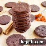 Chocolate cookies are scattered on a beige wooden surface, with a spoonful of cayenne pepper, cinnamon sticks, and a tall stack of cookies in the middle.