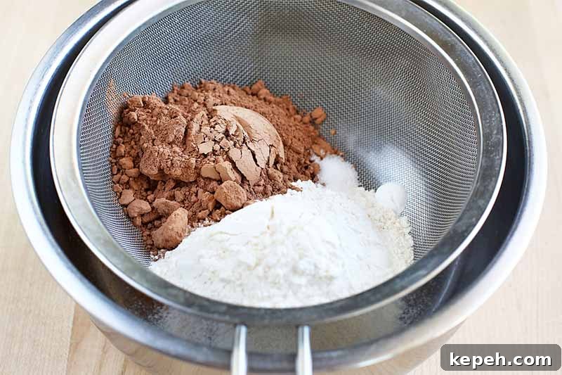 Spicy Mexican Hot Chocolate Cookies to Kick Off Baking Season 8 A metal mesh strainer inset in a stainless steel bowl is being used to sift white flour and cocoa powder, on a beige wooden background.