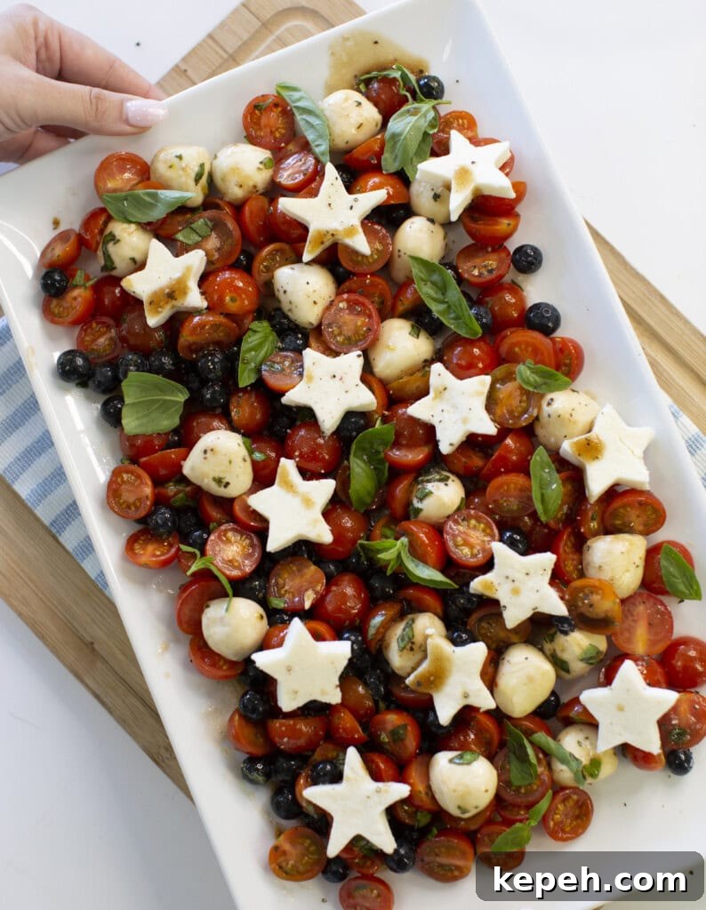 A platter of cherry tomatoes, black olives, mozzarella balls, and fresh basil, topped with star-shaped mozzarella slices, is shown from above. A hand holds the platter. The dish is placed on a striped cloth and wooden board.