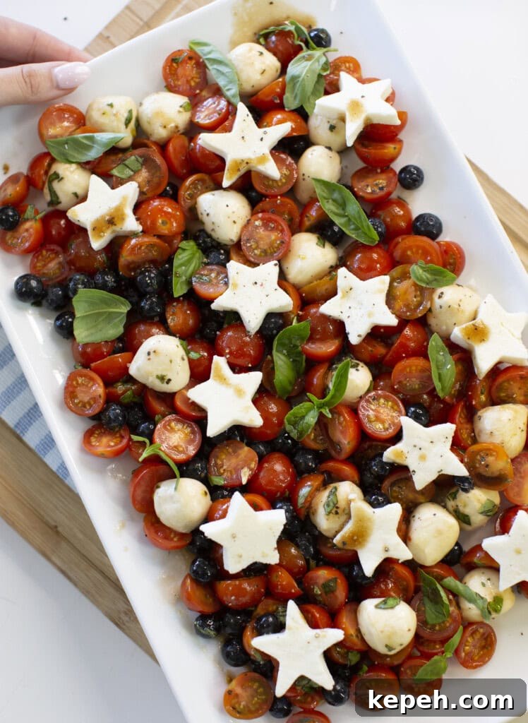 A rectangular platter with cherry tomatoes, blueberries, mozzarella balls, fresh basil, and star-shaped mozzarella slices, drizzled with dressing. A hand is holding the edge of the plate.