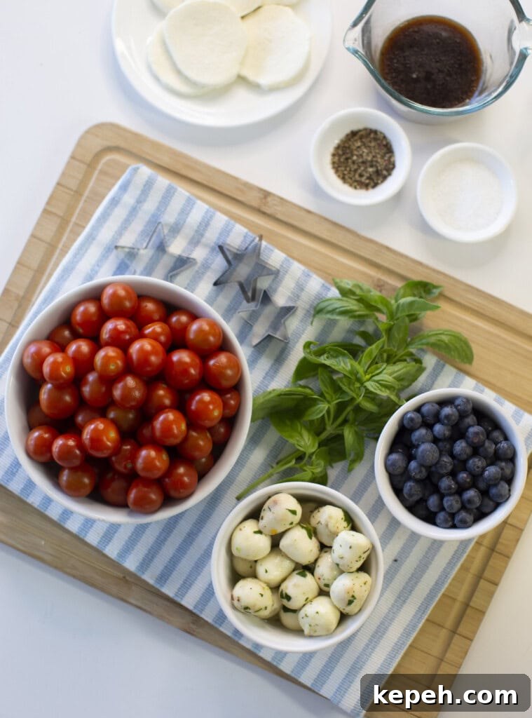 A cutting board with bowls of cherry tomatoes, blueberries, mozzarella balls, fresh basil, and small containers of spices and balsamic dressing, set on a blue-striped cloth. Slices of mozzarella are also visible.