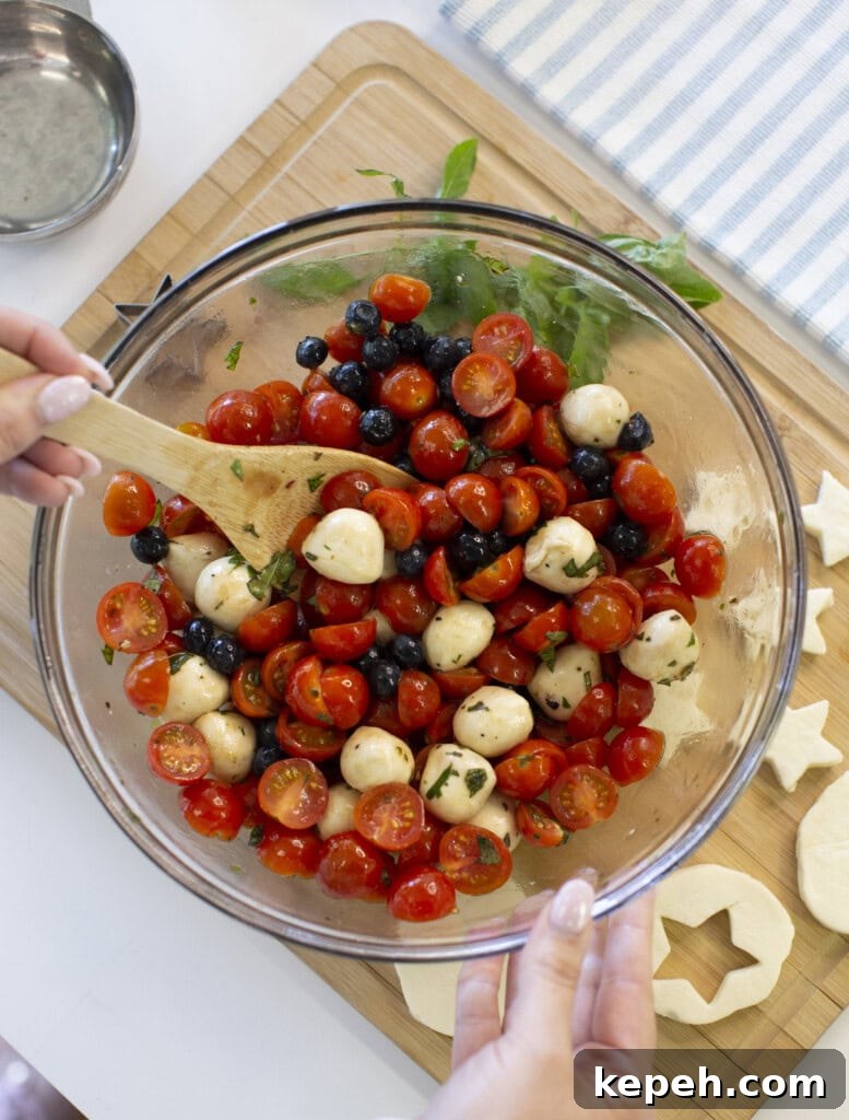 A person mixes a salad of cherry tomatoes, mozzarella balls, blueberries, and herbs in a glass bowl with a wooden spoon on a kitchen counter.