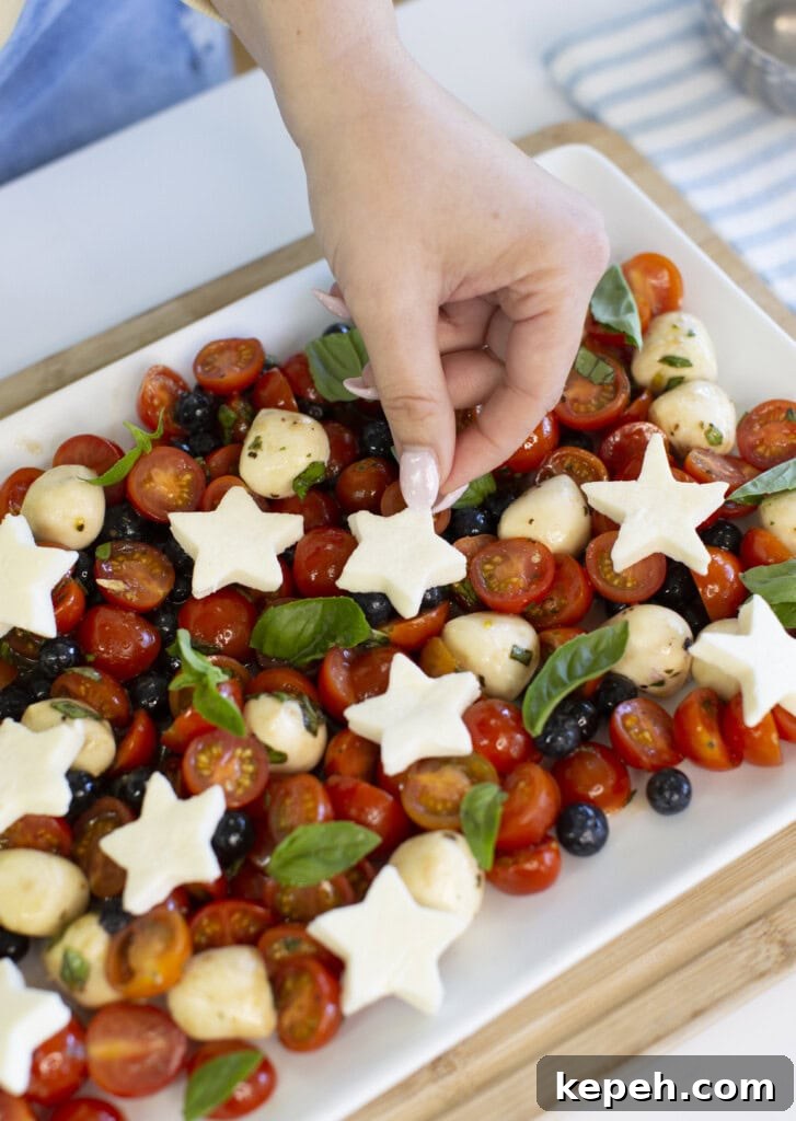 A hand places a star-shaped piece of cheese on a platter of cherry tomatoes, mozzarella balls, blueberries, and fresh basil.