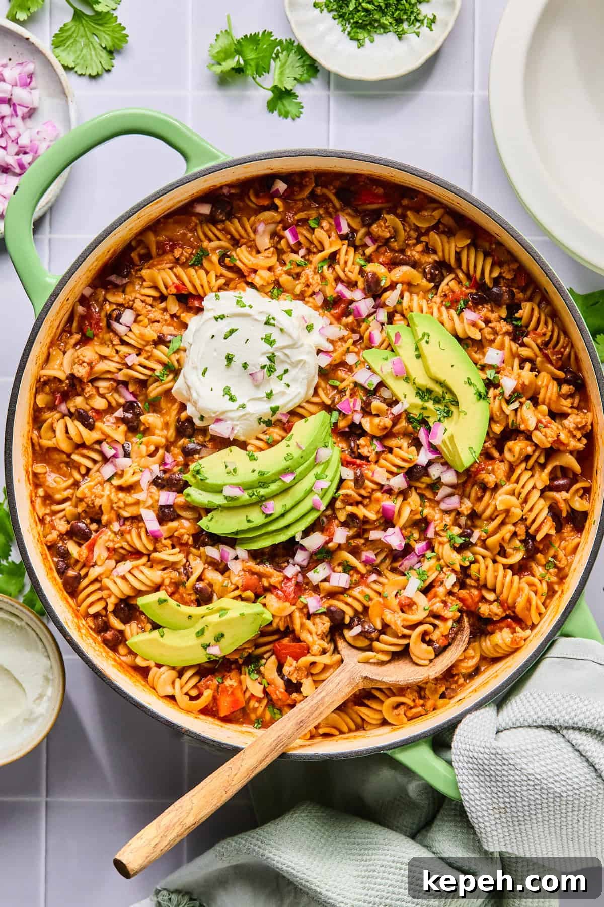 One pot taco pasta in the pot topped with sour cream, avocado, and cilantro with a wooden spoon.