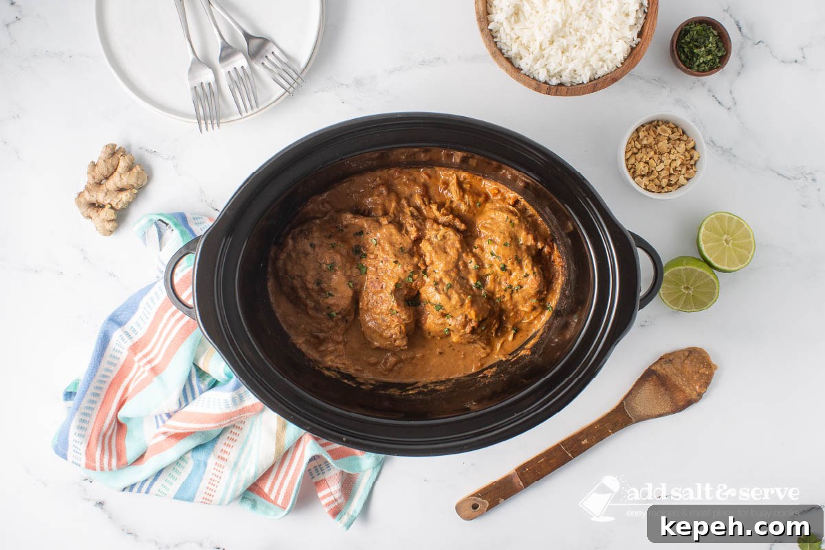 Slow Cooker Thai Peanut Chicken Recipe 7 Cooked Thai Peanut Chicken in a slow cooker surrounded by rice, chopped peanuts, cilantro, ginger, a halved lime, and a stack of empty plates with forks.